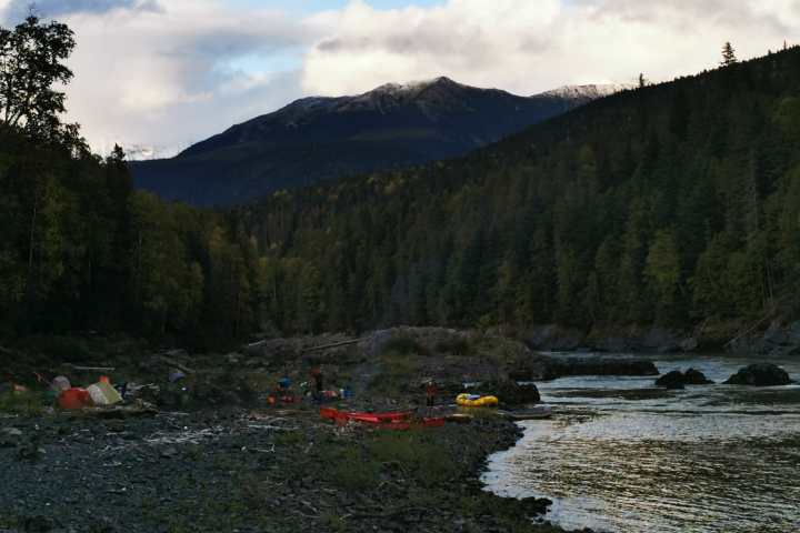Camp with kayaks on rocky riverbank in forested valley under cloudy sky.