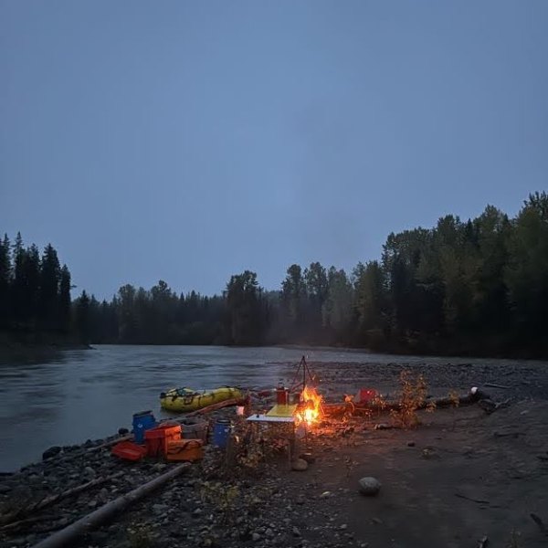 Campfire by riverbank with raft and trees at dusk.