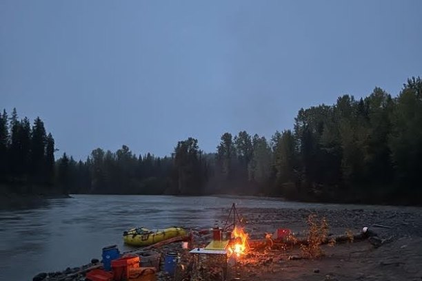 Campsite by river with campfire, supplies, and inflatable boat at dusk.