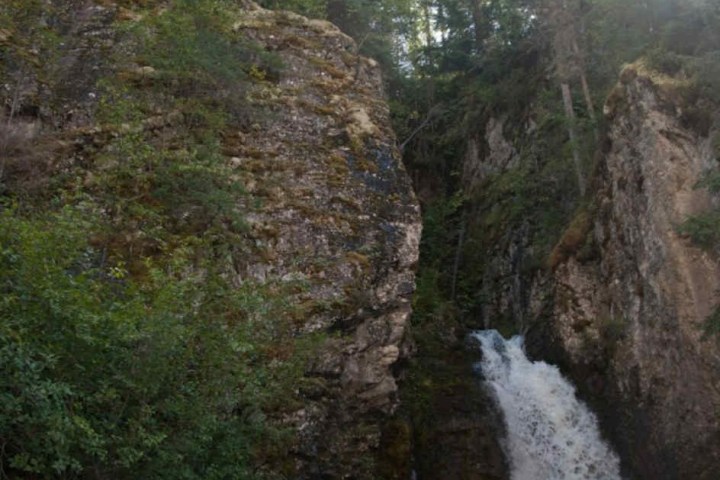 Tall rocky waterfall surrounded by trees and sunlight.