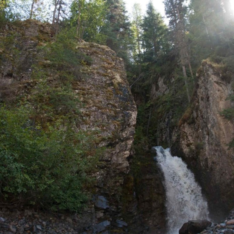 Tall rocky waterfall surrounded by trees and sunlight.