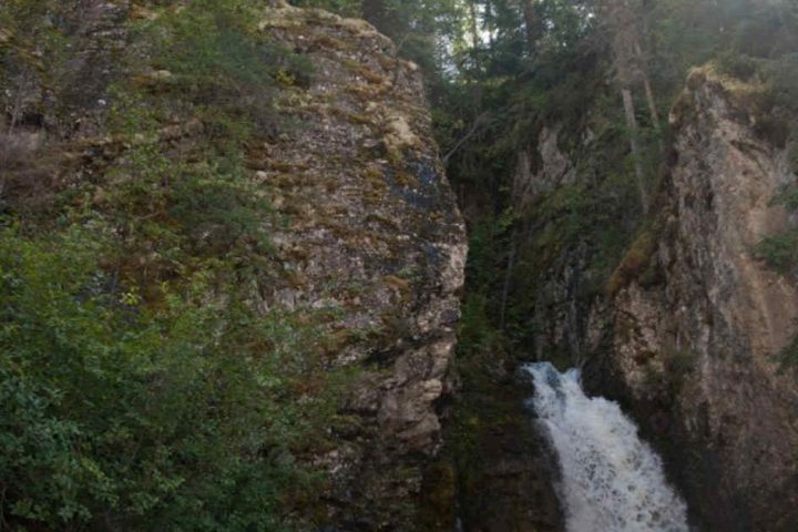 Waterfall cascading down rocky cliff surrounded by trees under a clear sky.