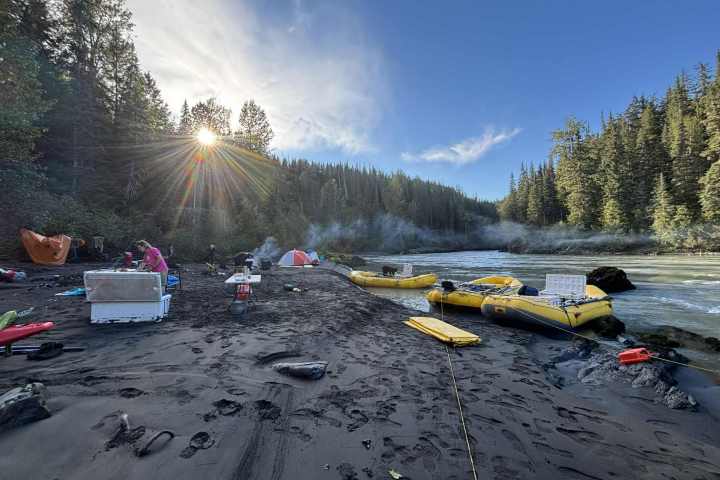 Riverbank campsite with tents, rafts, and sunrise through trees.