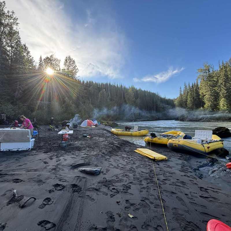 Riverbank campsite with tents, rafts, and sunrise through trees.