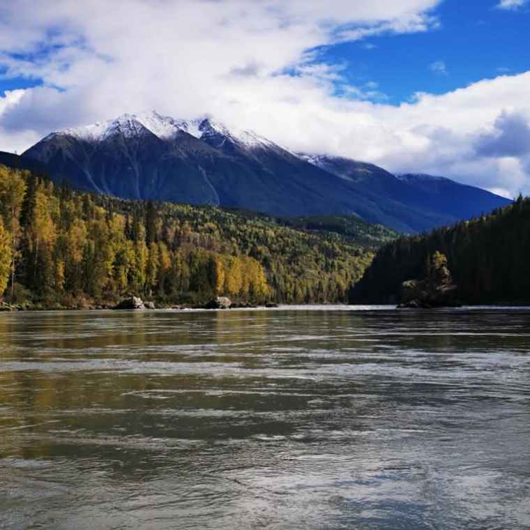 River flowing with mountains and trees in the background under a partly cloudy sky.