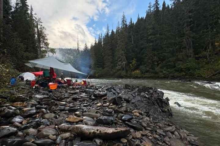 Riverside campsite with tarp, chairs, and trees under a cloudy sky.
