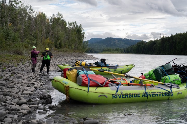Two people with gear beside three loaded inflatable rafts on a rocky riverbank.