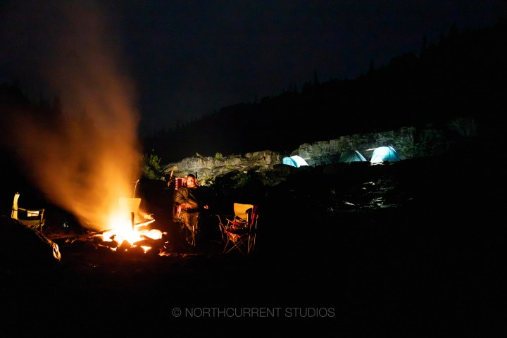 Night campsite with a fire, chairs, and glowing tents in the background.