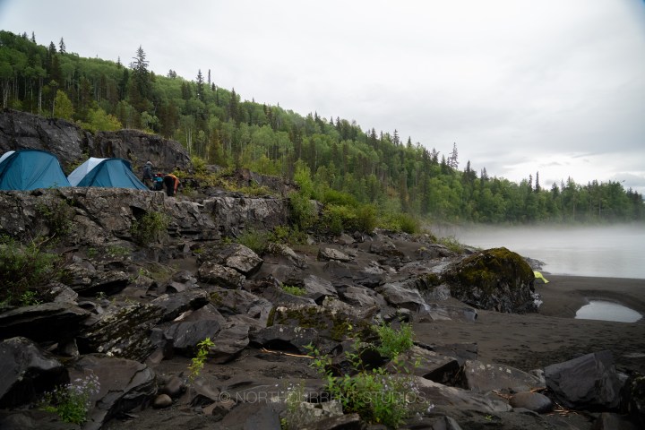 Tents on rocky terrain near a misty lake surrounded by lush green forest.