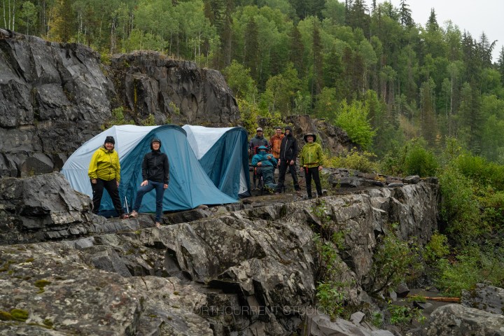 People in rain gear standing by a tent on rocky terrain in a forested area.