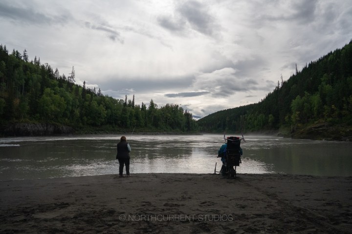 Two people by a calm river, surrounded by forested hills under a cloudy sky.