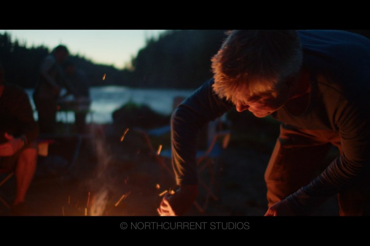 Person tending campfire by a lake at sunset, with sparks flying.