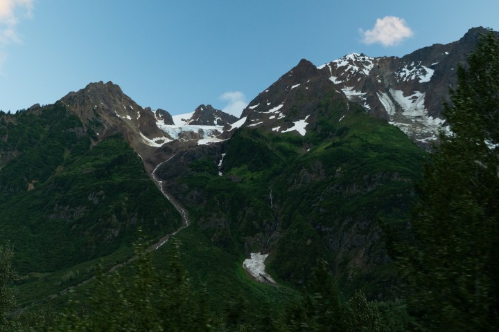 Mountain landscape with snow patches and green foliage under a clear blue sky.