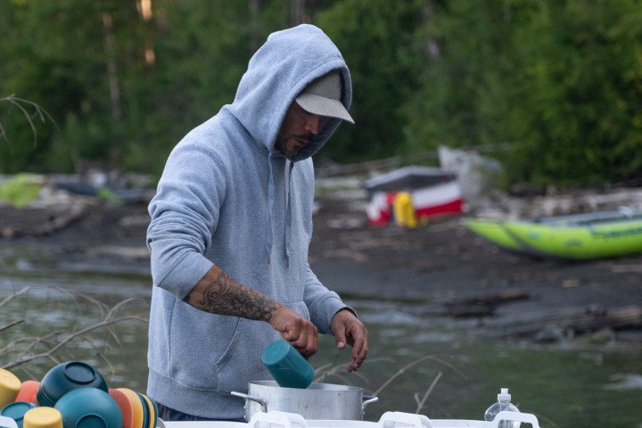 Person in hoodie preparing food at outdoor campsite with kayaks nearby.