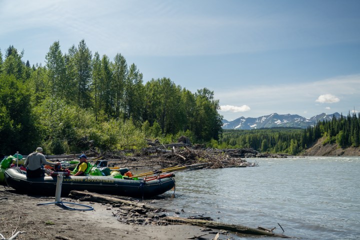 People preparing rafts by a riverbank with mountains in the background.