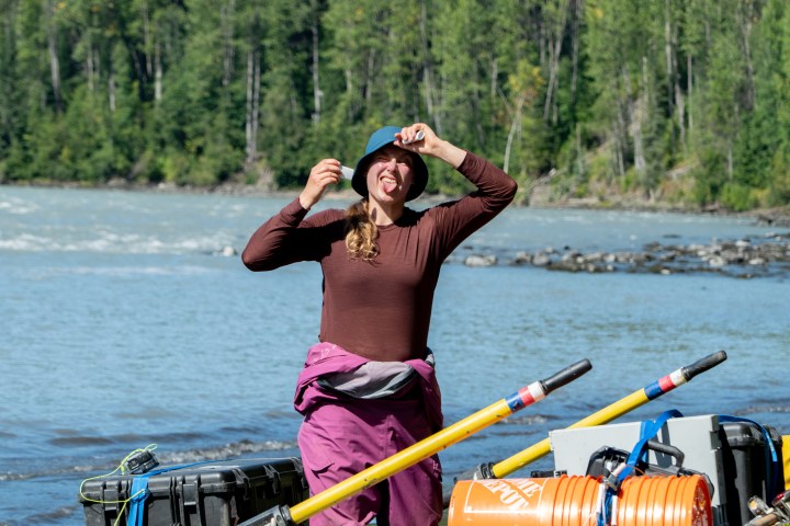 Person adjusting a hat on a raft near a river, trees in background.