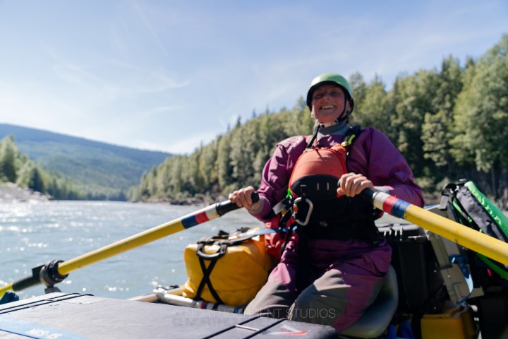 Person rowing a raft on a river, wearing a helmet and life jacket, surrounded by forested hills.