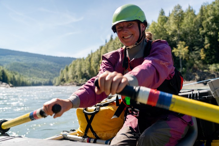 Smiling person in a helmet paddling a raft on a sunny river.
