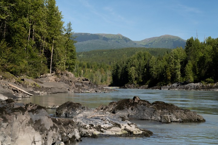 Calm river with rocky shore and forested hills in the background under blue sky.
