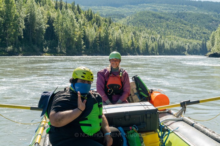 Two people in helmets and vests rafting on a river surrounded by forested hills.
