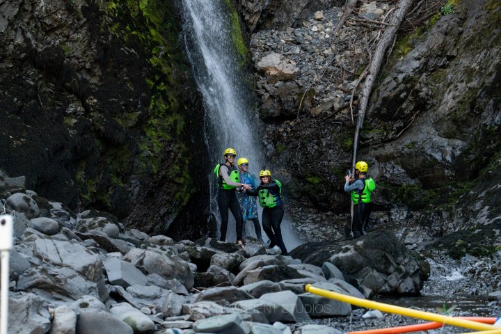 Four people in wetsuits and helmets standing near a waterfall.