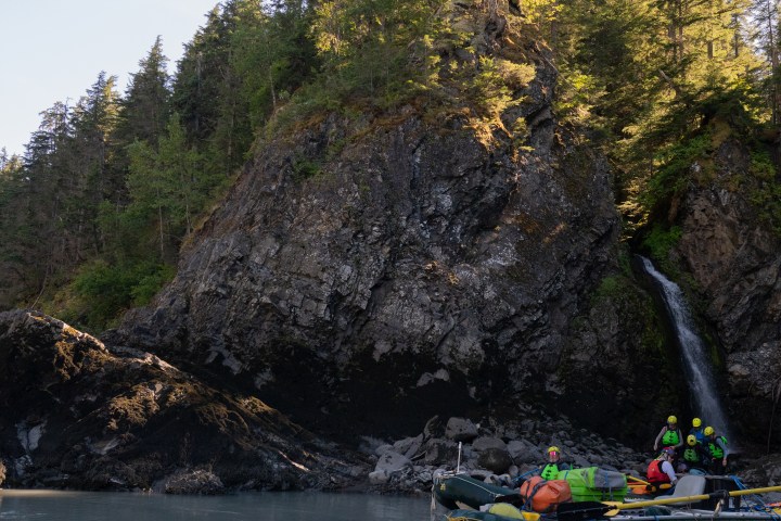 Rafting group near rocky cliff with waterfall and tall trees.