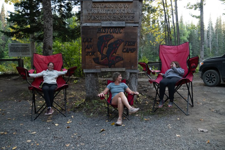 Three people sit in oversized chairs near a fishing site sign in a forested area.