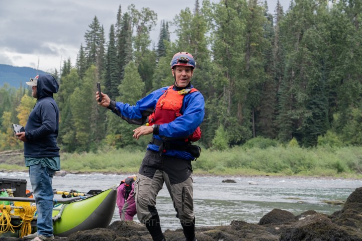 Man in blue life jacket posing on rocky riverbank with trees in background.