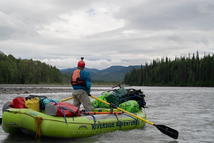 Person on a raft with gear, navigating a wide river surrounded by trees.